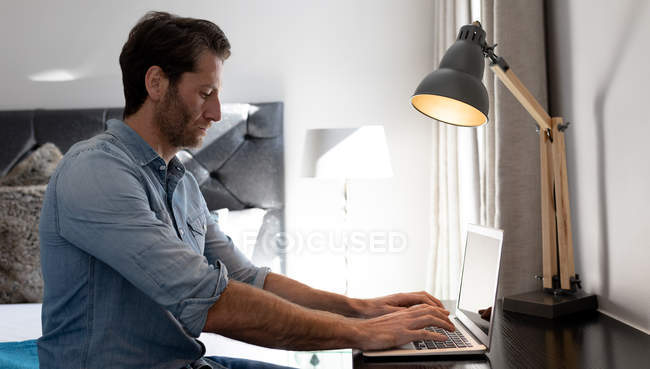 Vista lateral de un joven caucásico que se relaja de vacaciones en una habitación de hotel con un ordenador portátil - foto de stock