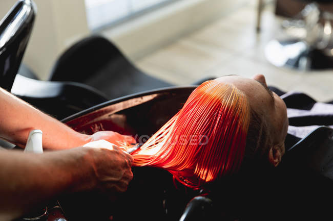 Visão traseira close-up de um cabeleireiro masculino caucasiano de meia idade e uma jovem mulher caucasiana com seu cabelo colorido vermelho brilhante, lavado e penteado em um salão de cabeleireiro — Fotografia de Stock