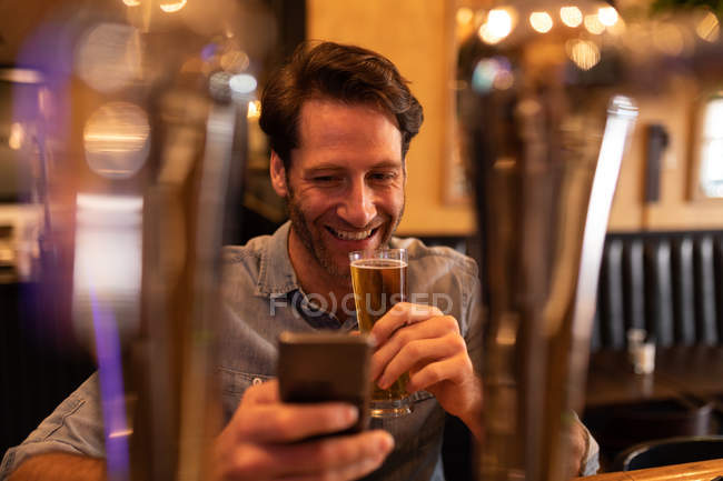 Вид на молоду Кавказьку людину розслабляє на відпочинку в барі, питне пиво і за допомогою смартфона — стокове фото