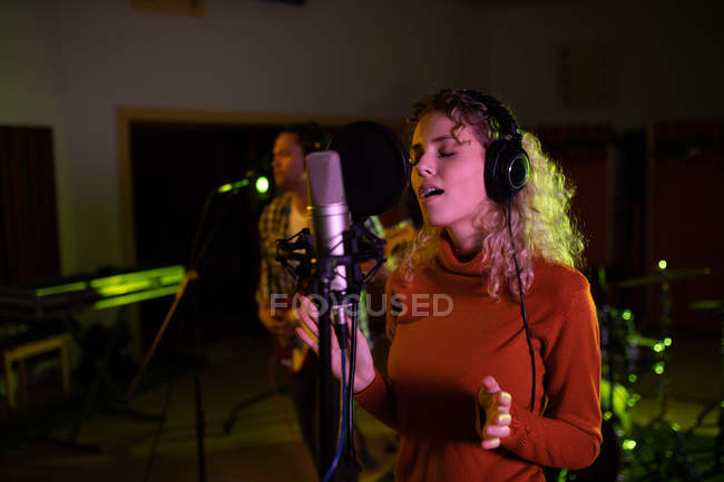 Vista frontal de uma jovem cantora caucasiana usando fones de ouvido cantando na frente de um microfone em um estúdio de gravação, gesticulando e com os olhos fechados — Fotografia de Stock