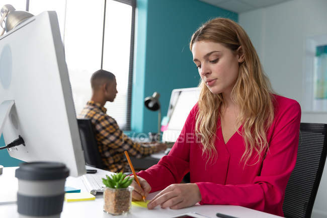 Front view close up of a young Caucasian woman sitting at a desk writing a note, with her male colleague sitting and working at a computer in the background, at the modern office of a creative business — Stock Photo