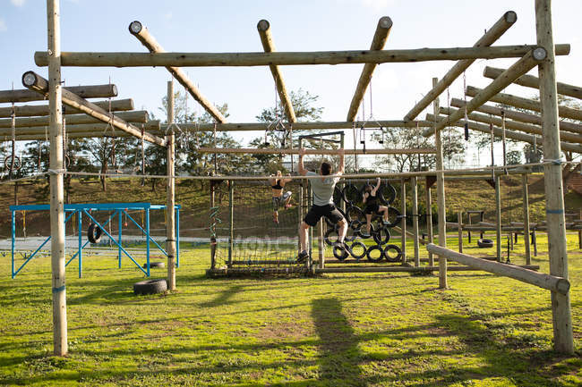 Rear view of a young Caucasian man hanging on a climbing frame at an outdoor gym during a bootcamp training session, with other participants in the background — Stock Photo