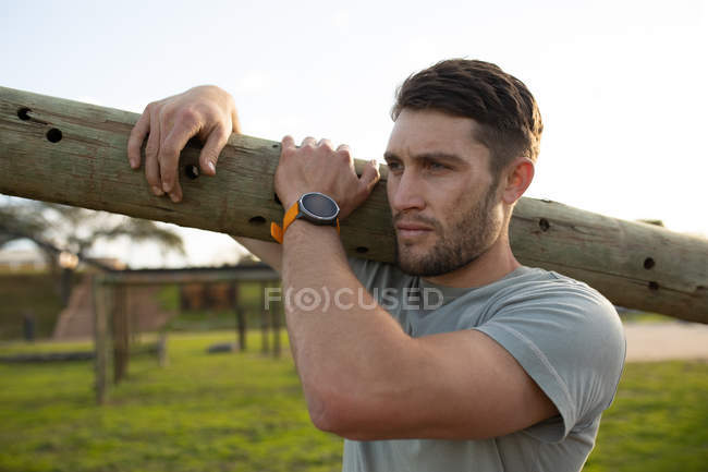 Vista lateral de un joven caucásico llevando un tronco en su hombro en un gimnasio al aire libre durante una sesión de entrenamiento de bootcamp - foto de stock