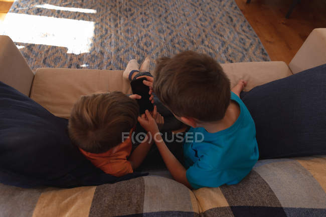 Вид сверху на двух кавказских мальчиков, сидящих на диване и использующих смартфон в гостиной — стоковое фото