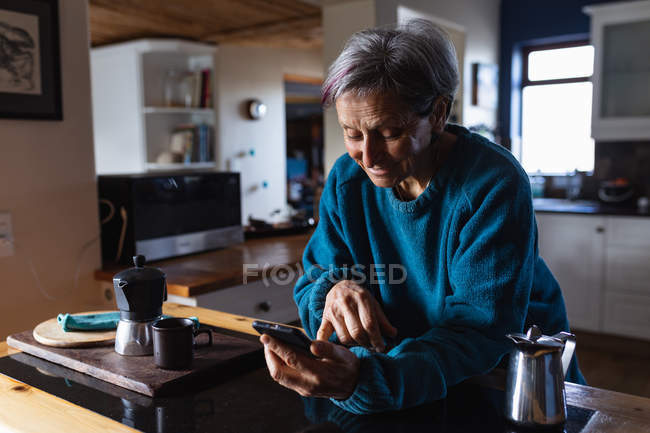 Вид збоку крупним планом старший кавказька жінка на кухні за допомогою смартфона з кухонних шаф у фоновому режимі — стокове фото
