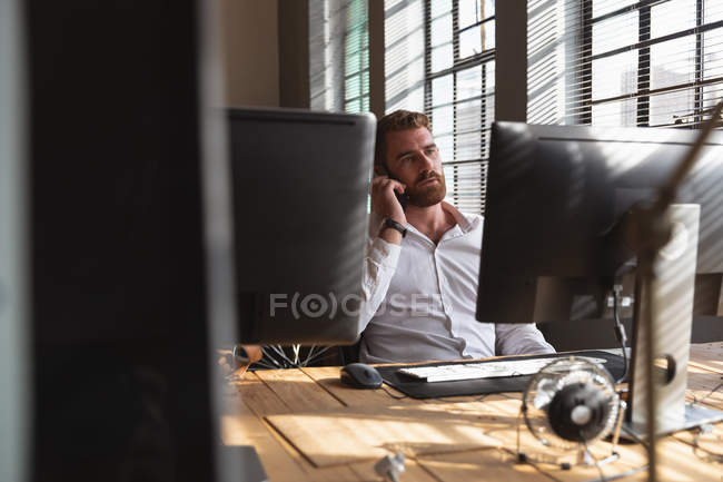 Vista frontale da vicino di un giovane caucasico seduto a una scrivania che parla su uno smartphone in un ufficio creativo, visto tra gli schermi del computer — Foto stock