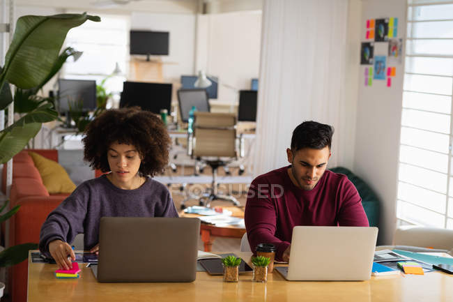 Вид спереду молодого змішаного чоловіка і молодої змішаної раси жінки, що сидить за столом, використовуючи ноутбук комп'ютерів у творчому офісі — стокове фото