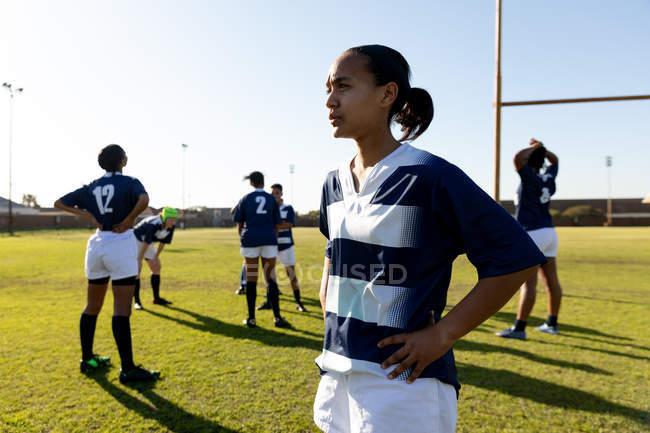 Front view of a young adult mixed race female rugby player standing on a rugby pitch with her hands on her hips looking away, with her teammates stretching in the background — Stock Photo