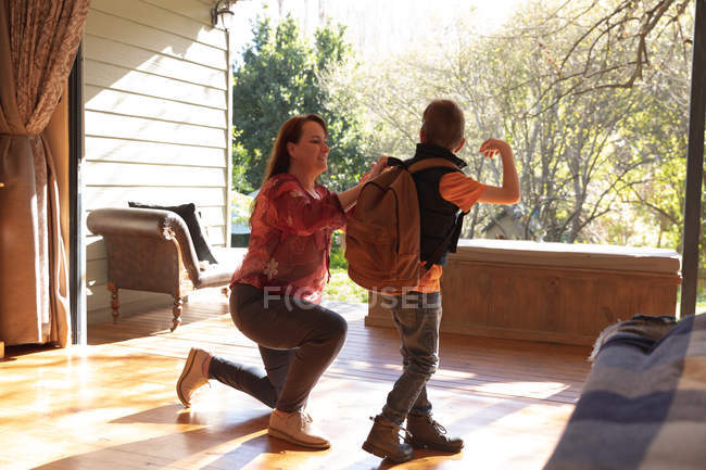 Vista lateral de uma mulher caucasiana de meia idade ajoelhada ao lado de seu filho pré-adolescente e tirando sua mochila — Fotografia de Stock