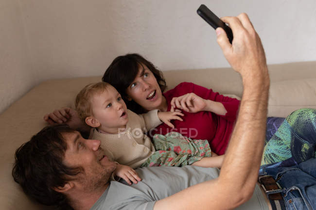 Vista lateral de cerca de un joven padre y madre caucásicos acostados en un sofá con su bebé y tomando una selfie - foto de stock