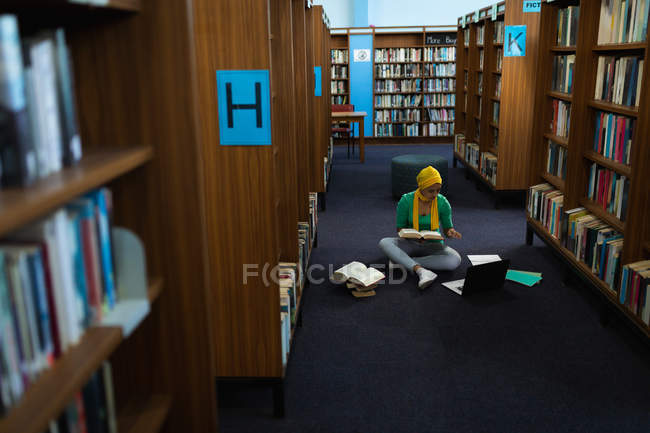 Vorderseite einer jungen asiatischen Studentin mit Turban, die ein Buch in der Hand hält, einen Laptop benutzt und in einer Bibliothek studiert — Stockfoto
