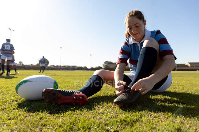 Vue de face gros plan d'une jeune joueuse de rugby blanche adulte assise et attachant sa botte sur un terrain de rugby avec la balle à côté d'elle et ses coéquipiers en arrière-plan — Photo de stock