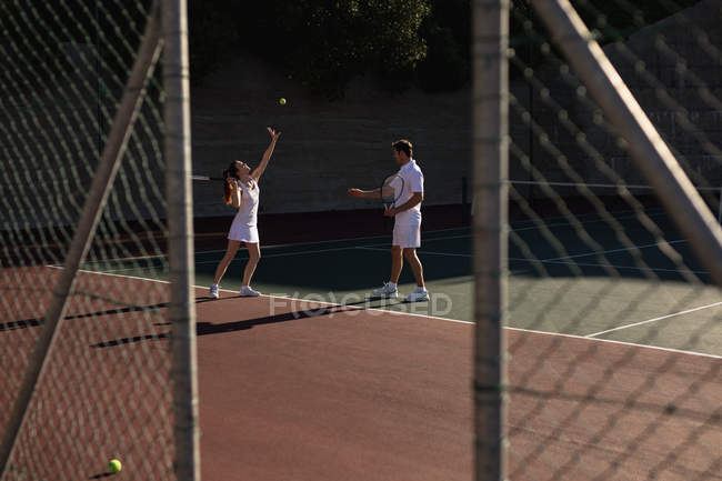 Вид сбоку на молодую кавказку и мужчину, играющего в теннис в солнечный день, женщину, готовящуюся служить, и жест мужчины, увиденный через забор — стоковое фото