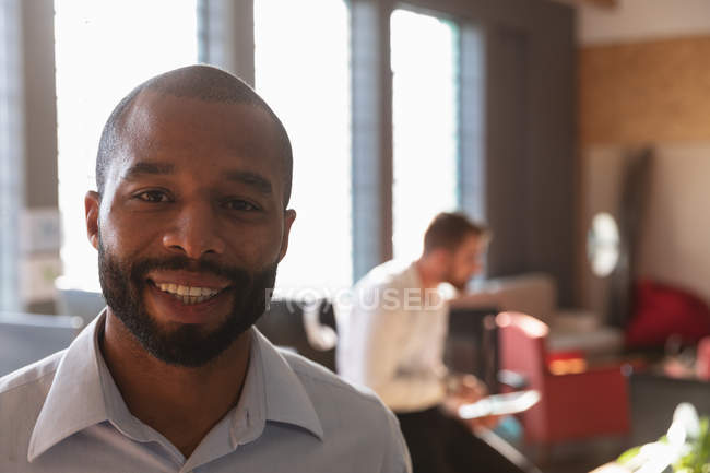 Portrait close up of a young African American man with short hair and a beard looking to camera smiling in a creative office with colleague working in the background — Stock Photo