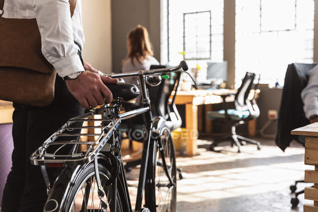 Visão lateral na metade da seção de um jovem caucasiano que leva uma bicicleta para um escritório criativo, com uma colega sentada em uma mesa trabalhando em segundo plano — Fotografia de Stock