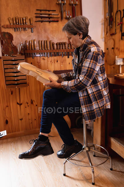 Side view of a senior Caucasian female luthier holding the body of a violin in her workshop, with tools hanging up on the wall in the background — Stock Photo