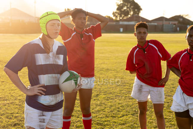 Frontansicht einer Gruppe junger erwachsener multiethnischer Rugbyspielerinnen, die nach einem Rugbyspiel entspannt auf einem Rugbyfeld stehen, ein kaukasischer Spieler mit Kopfschutz hält den Ball — Stockfoto