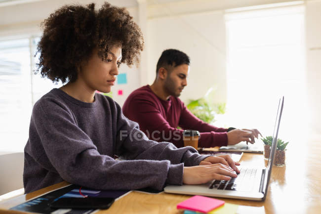 Vue de côté gros plan d'un jeune homme de race mixte et d'une jeune femme de race mixte assis à un bureau à l'aide d'ordinateurs portables dans un bureau créatif — Photo de stock