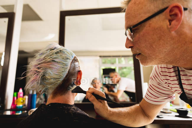 Vue de côté gros plan d'un homme caucasien d'âge moyen coiffeur et une jeune femme caucasienne ayant ses cheveux colorés dans un salon de coiffure, reflétée dans un miroir — Photo de stock