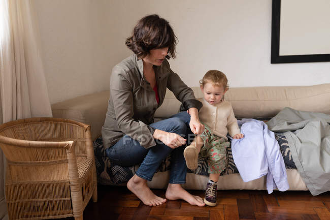 Front view of a young Caucasian mother getting her baby dressed, sitting on a couch — Stock Photo