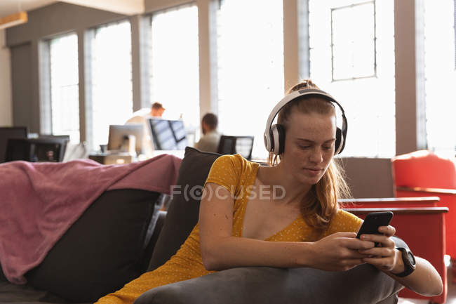 Vista frontal de cerca de una joven mujer caucásica sentada con auriculares y usando un teléfono inteligente en la sala de estar de una oficina creativa con colegas trabajando en escritorios y computadoras en el fondo - foto de stock