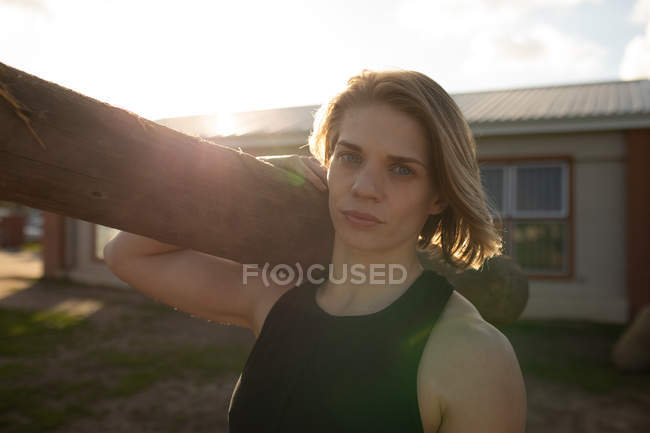 Portrait close up of a young Caucasian woman carrying a log on her shoulder at an outdoor gym during a bootcamp training session — Stock Photo