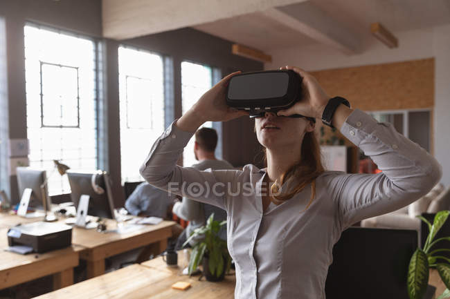 Vista frontal de cerca de una joven mujer caucásica con un casco VR de pie y mirando hacia arriba en una oficina creativa, con colegas trabajando en segundo plano - foto de stock
