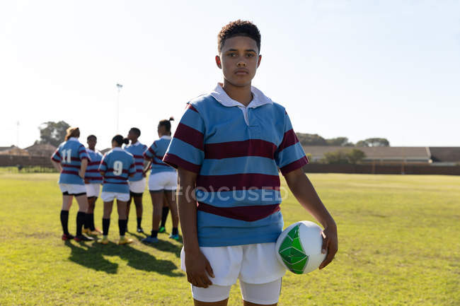 Portrait d'une jeune joueuse de rugby mixte adulte debout sur un terrain de rugby tenant une balle de rugby regardant vers la caméra, avec ses coéquipières parlant ensemble en arrière-plan — Photo de stock