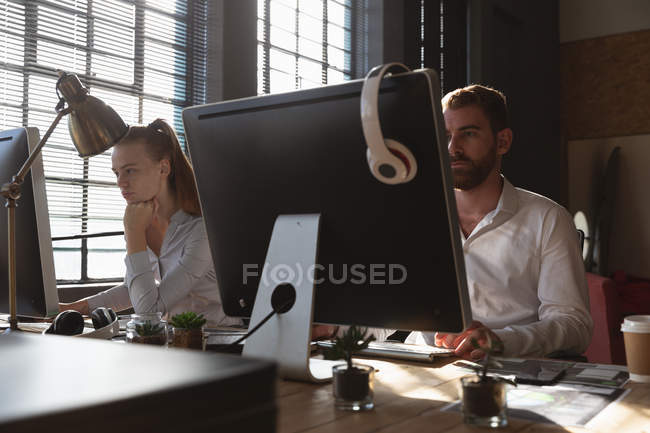 Вид спереди на молодого кавказца, женщину и мужчину, сидящих за столом с компьютерами в творческом офисе — стоковое фото