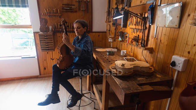 Frontansicht einer älteren kaukasischen Geigenbauerin, die in ihrer Werkstatt eine Geige hält, mit einem Tablet-Computer auf einer Werkbank und Werkzeug an der Wand im Hintergrund — Stockfoto