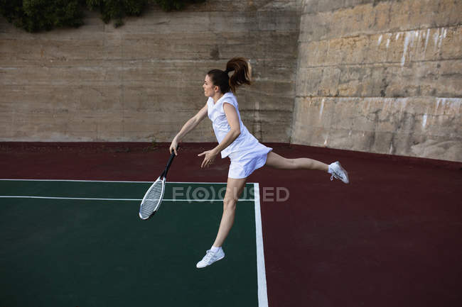 Seitenansicht einer jungen kaukasischen Frau, die Tennis spielt, einen Ball zurückbringt und mit einer Wand hinter sich springt — Stockfoto