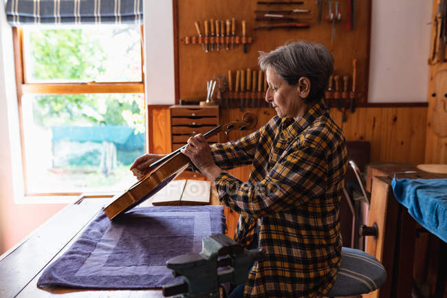 Vista laterale di una liutaia caucasica anziana che lavora su un violino nel suo laboratorio con strumenti appesi al muro sullo sfondo — Foto stock
