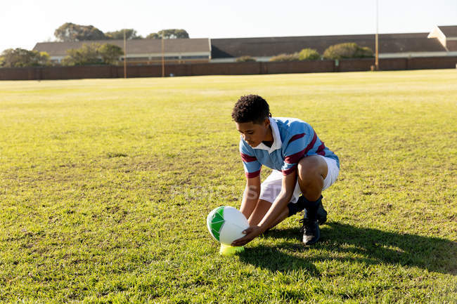 Вид спереди на молодую взрослую смешанную расу регбистку, стоящую на коленях на поле для регби и ставящую мяч на футболку для удара по месту — стоковое фото