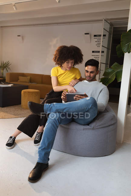 Вид спереду крупним планом молодий чоловік змішаної раси і молода змішана жінка, що сидить на мішку з квасолею, використовуючи планшетний комп'ютер і обговорюючи в творчому офісі — стокове фото