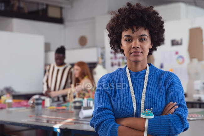 Retrato de cerca de una joven estudiante de moda de raza mixta mirando directamente a la cámara en un estudio de la universidad de moda, con estudiantes trabajando en el fondo - foto de stock
