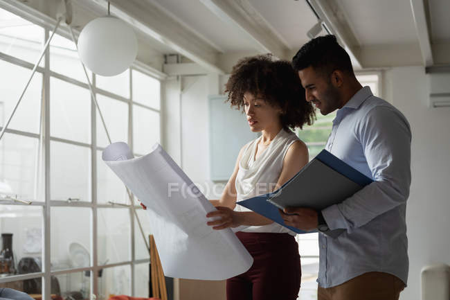 Vista laterale da vicino di un giovane uomo di razza mista e una giovane donna di razza mista in piedi e controllando un disegno architettonico in un ufficio creativo — Foto stock
