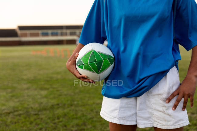Передня частина жіночого регбіста, що стоїть на спортивному полі, тримає м'яч регбі під час тренувальної сесії — стокове фото