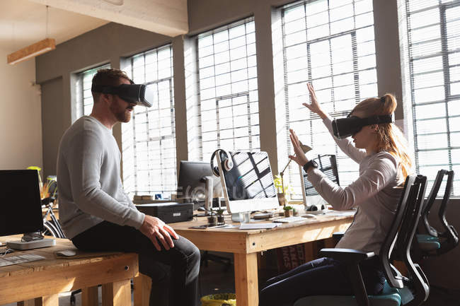 Side view close up of a young Caucasian man sitting on a desk and a young Caucasian woman sitting on a chair both wearing a VR headsets in a creative office — Stock Photo