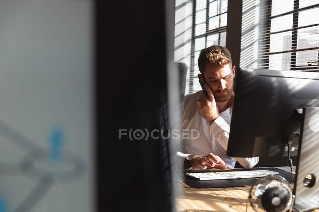 Nahaufnahme eines jungen kaukasischen Mannes, der an einem Schreibtisch sitzt, mit einem Smartphone spricht und einen Computer in einem kreativen Büro benutzt, gesehen zwischen Computerbildschirmen — Stockfoto