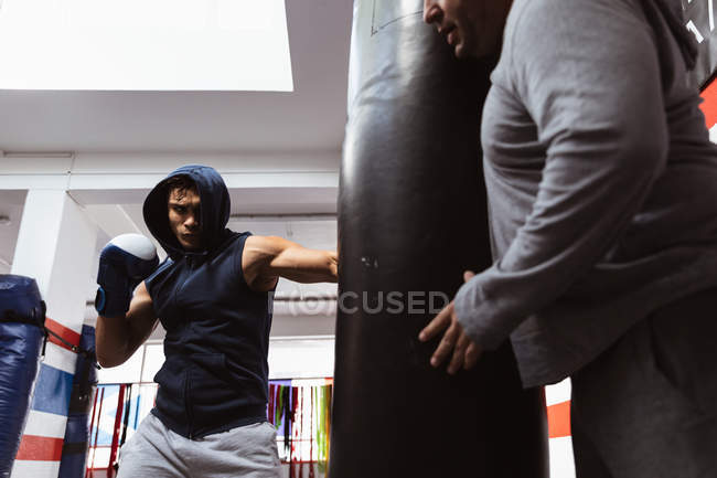 Vista frontal close-up de um jovem lutador de boxe misto perfurando um saco de pancada segurado por um treinador masculino caucasiano de meia idade em um ginásio de boxe — Fotografia de Stock