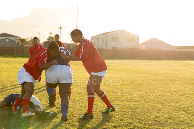Vista lateral de dos jugadoras de rugby de raza mixta adultas jóvenes que abordan a un jugador del equipo contrario durante un partido de rugby, con otros jugadores en el fondo y uno tumbado en el suelo - foto de stock