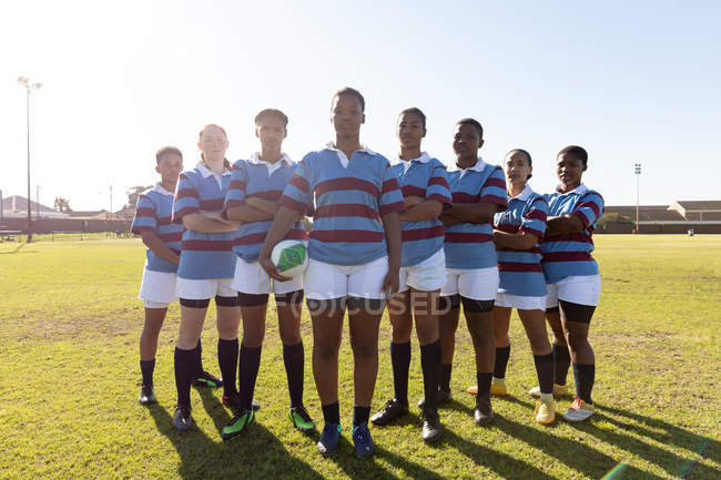 Retrato de un equipo de jóvenes jugadoras de rugby multiétnicas adultas en formación en un campo de rugby con los brazos cruzados, mirando a la cámara en un campo de rugby, la mujer en la parte delantera sostiene una pelota de rugby - foto de stock