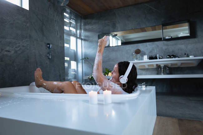 Side view of a young Caucasian brunette woman sitting in a foam bath wearing headphones, listening to music and singing along with her eyes closed and arm raised in the air — Stock Photo