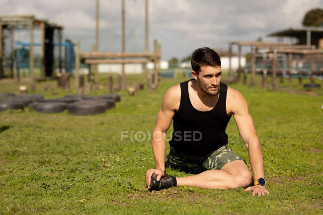 Vista frontal de un joven caucásico sentado en la hierba y estirándose en un gimnasio al aire libre antes de una sesión de entrenamiento de bootcamp - foto de stock
