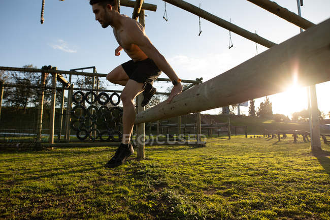 Vista lateral de um jovem caucasiano saltando sobre um obstáculo em um ginásio ao ar livre durante uma sessão de treinamento de bootcamp — Fotografia de Stock