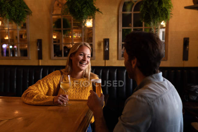 Вид спереди на счастливую молодую кавказскую пару, отдыхающую вместе в баре, пьющую пиво, вино и улыбающуюся — стоковое фото