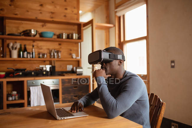 Вид збоку крупним планом молодого афроамериканського чоловіка, що носить гарнітуру VR, використовуючи ноутбук, сидячи за кухонним столом вдома — стокове фото