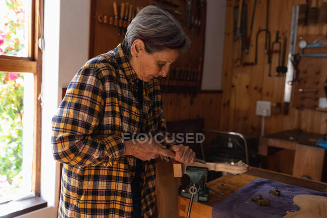 Vista lateral de uma luthier feminina caucasiana sênior trabalhando no pergaminho de um violino em sua oficina com ferramentas penduradas na parede no fundo — Fotografia de Stock
