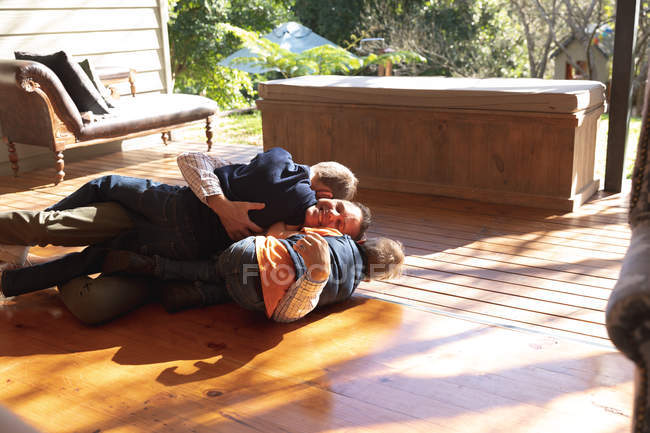 Vista frontal de un hombre caucásico de mediana edad acostado en el suelo abrazándose con sus dos hijos preadolescentes después de llegar a casa - foto de stock