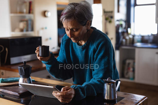 Вид збоку старший кавказька жінка в кухні пити каву і за допомогою планшетного комп'ютера з кухонних шаф у фоновому режимі — стокове фото